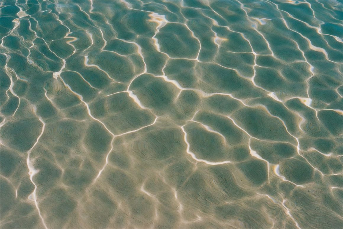 Sunlight refracting through shallow seawater into rippling bands of light on pale sand